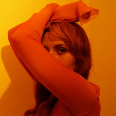 Alex peeks at camera from behind her arms, bathed in orange light
