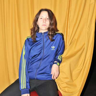 monibi wearing a blue and yellow adidas jacket stares boldly at the camera in front of a yellow curtain