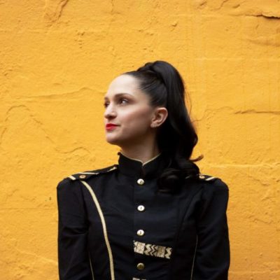 Nathalie wears a military style jacket, bright red lipstick and a high ponytail in front of a yellow wall
