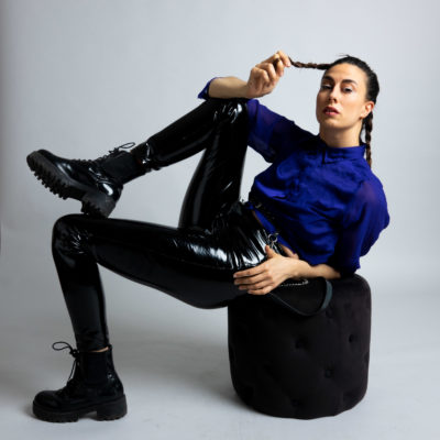 Anna poses on a stool wearing combat boots, black latex tights, a blue blouse and braids